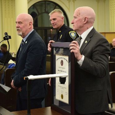 Three white men at City Commission speaking podium, with framed certificate
