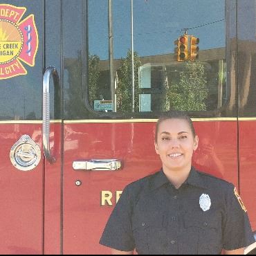White woman with dark hair in dark blue uniform, standing in front of red/black fire vehicle.